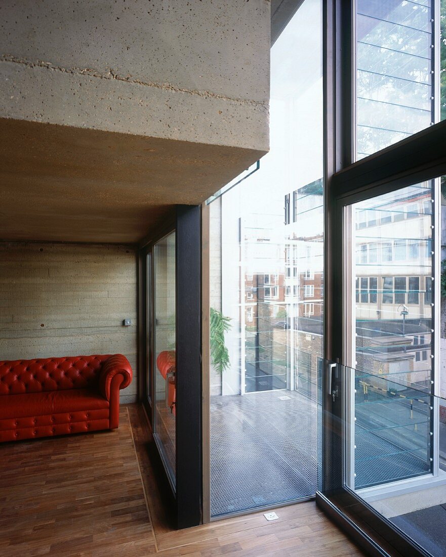 Ausschnitt eines Wohnraumes in Betonausführung mit ziegelrotem Ledersofa und Blick durch Fenster auf Balkon