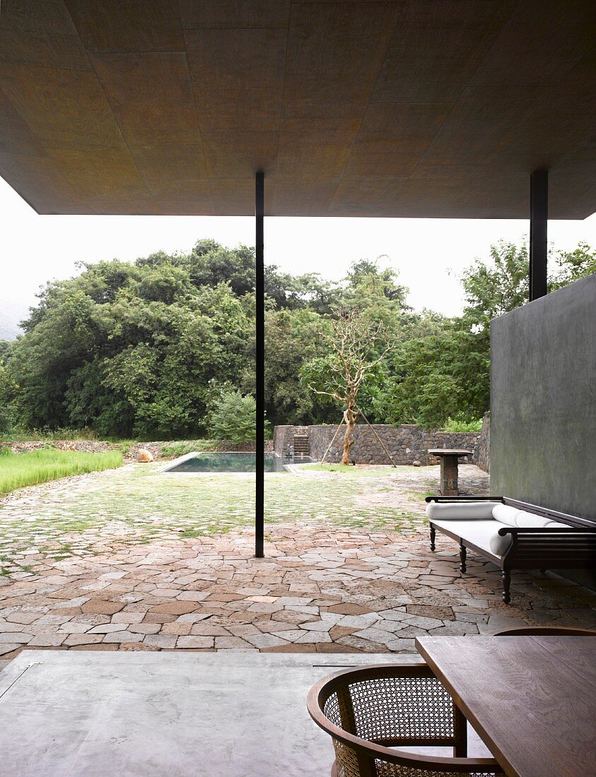 Antike indische Sitzbank vor Betonwand auf moderner überdachter Terrasse und Garten mit Pool