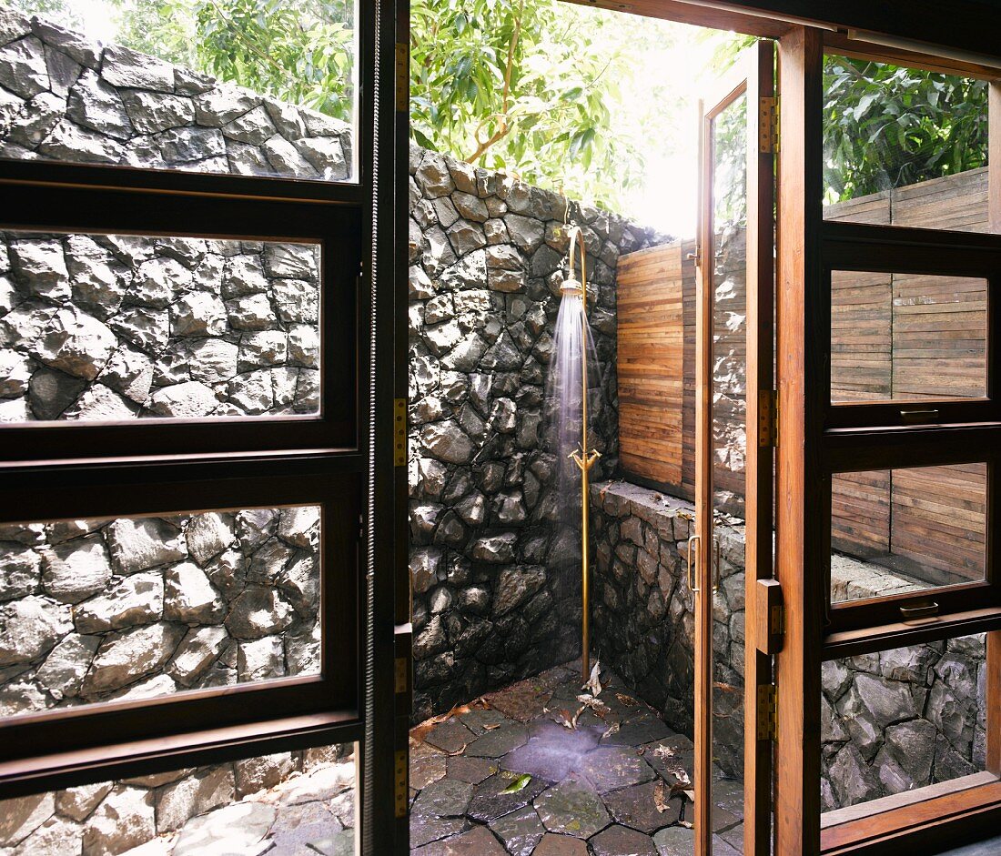 View through open terrace door of vintage brass outdoor shower in corner by stone wall