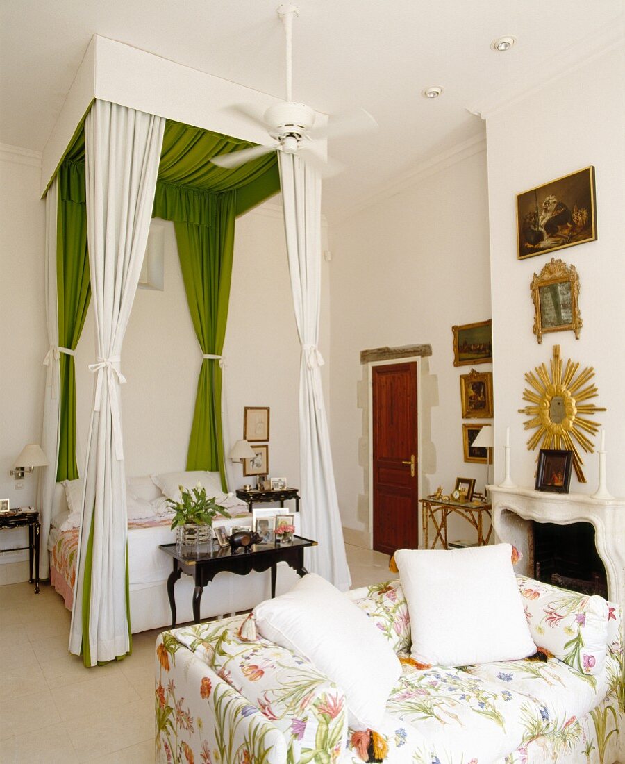 A bedroom with a floral patterned sofa in front of a four poster bed with a canopy made of green and white material