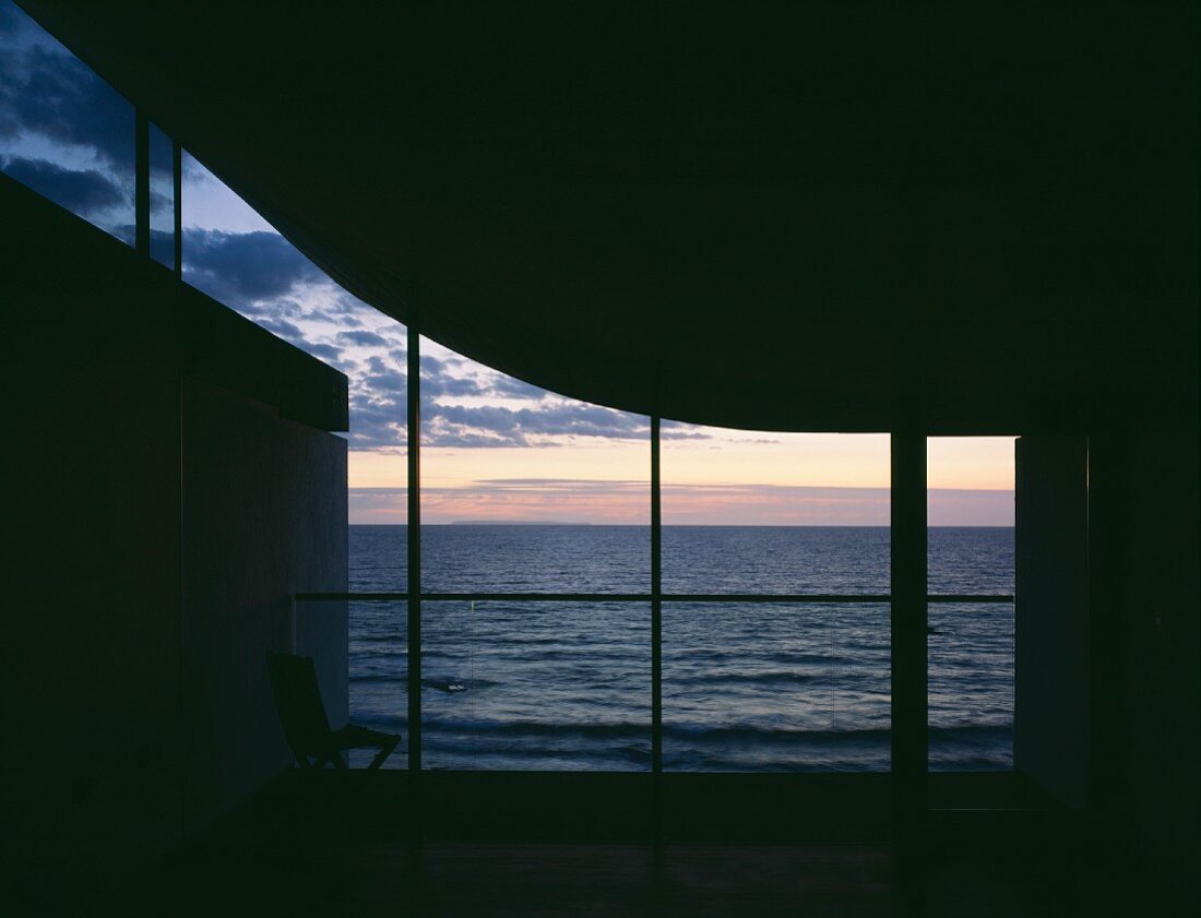 Skulpturaler, unbeleuchteter Innenraum mit Blick durch Glasfassade auf Abendhimmel über dem Meer