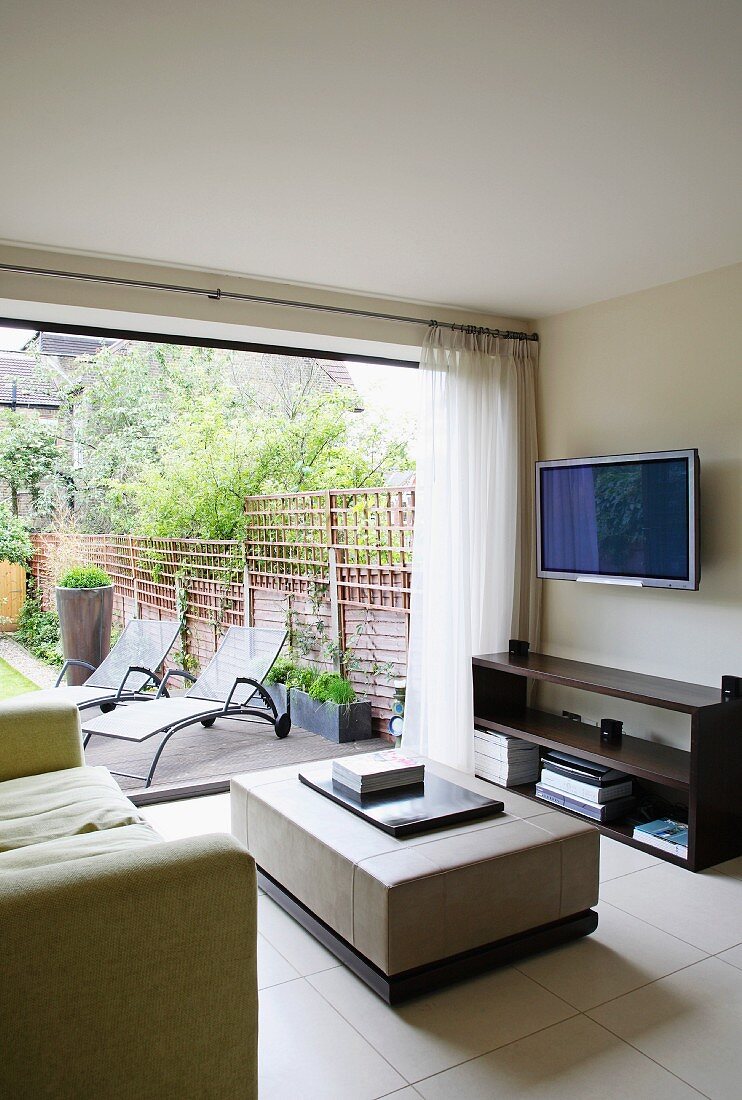 Modern TV area with view through open window wall to metal sun loungers in garden