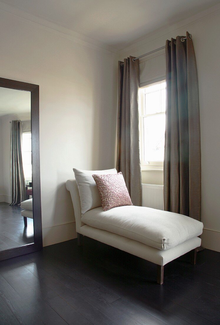 Wood-framed mirror and white chaise longue in front of window with sand-coloured curtains