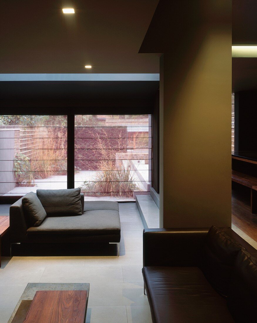 Open-plan living room in dark shades with various sofas in a sunken seating area and view into a garden through large windows