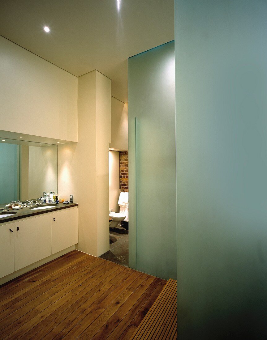 Large mirror above built-in washstands in modern bathroom with frosted glass walls and untreated wooden floorboards