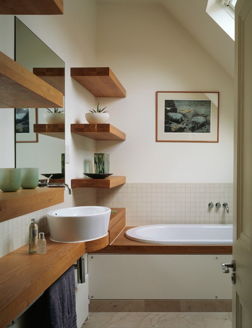 Wooden washstand, shelves and bathtub rim in small bathroom with skylight in sloping ceiling