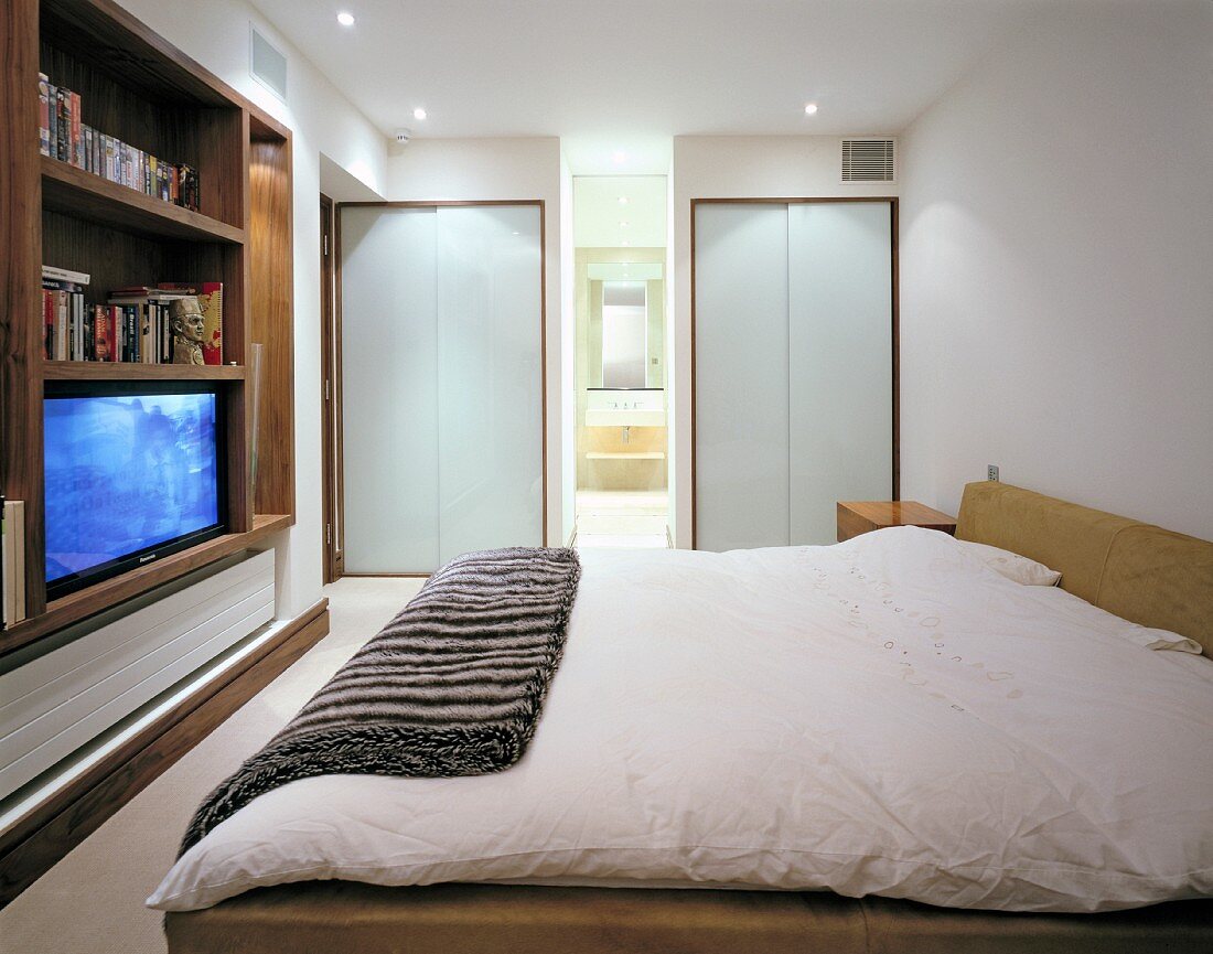Schlafzimmer Schrankwand mit laufendem Fernseher gegenüber Doppelbett in modernem Schlafzimmer mit Bad ensuite