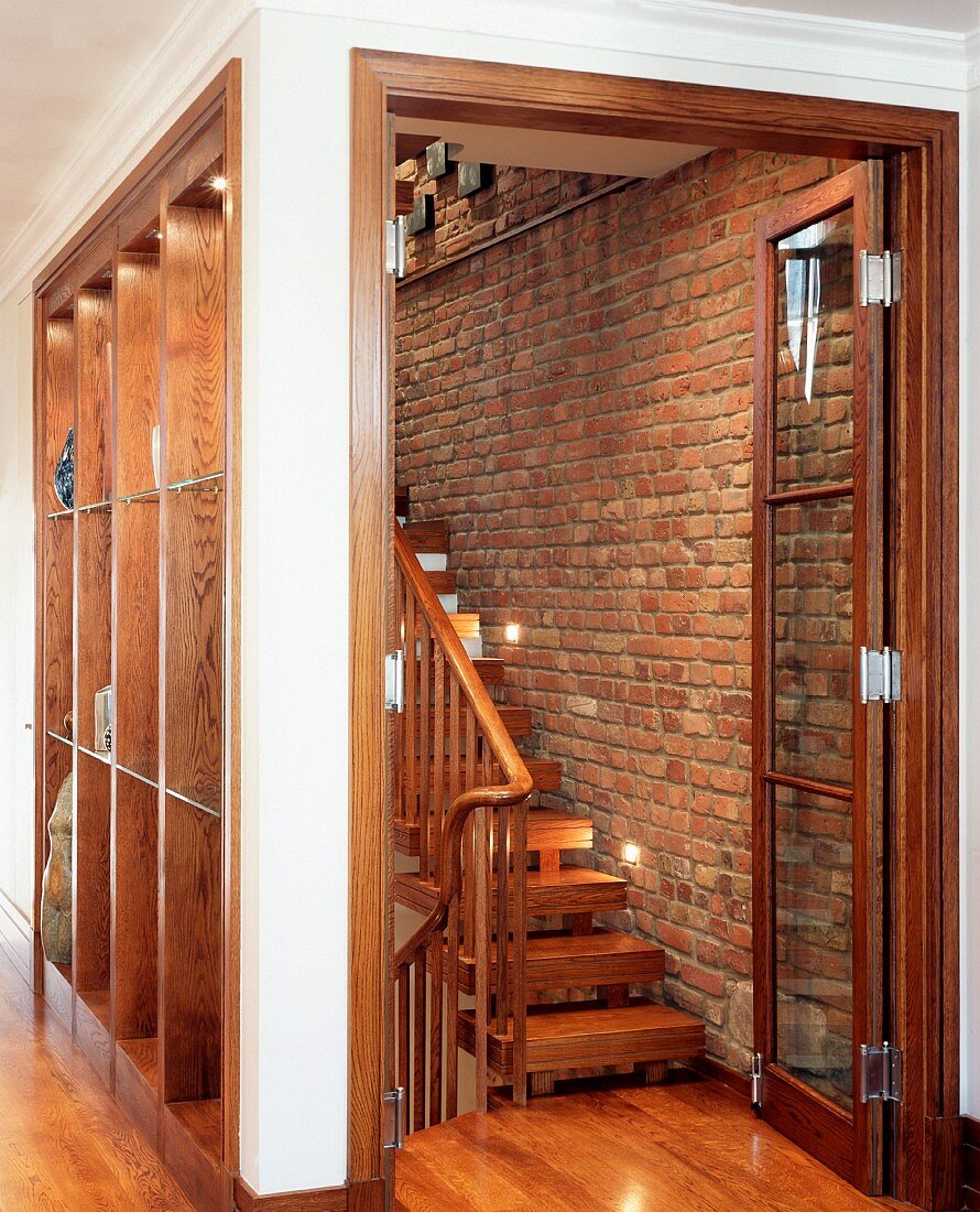 Vom Wohnraum abgetrennter Treppenraum mit Glastür und Holztreppe vor Backsteinwand