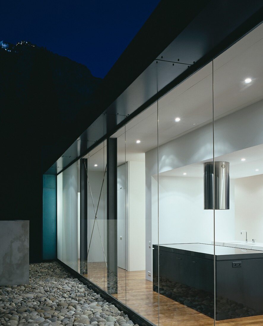 Evening on a terrace with a gravel floor and a view into an illuminated, open-plan, designer kitchen
