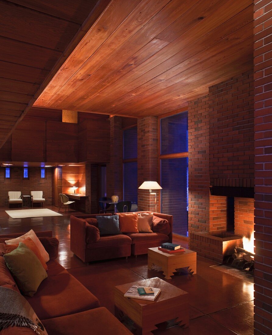 Evening atmosphere in modernist house with wooden ceiling and sofas next to fireplace in brick wall