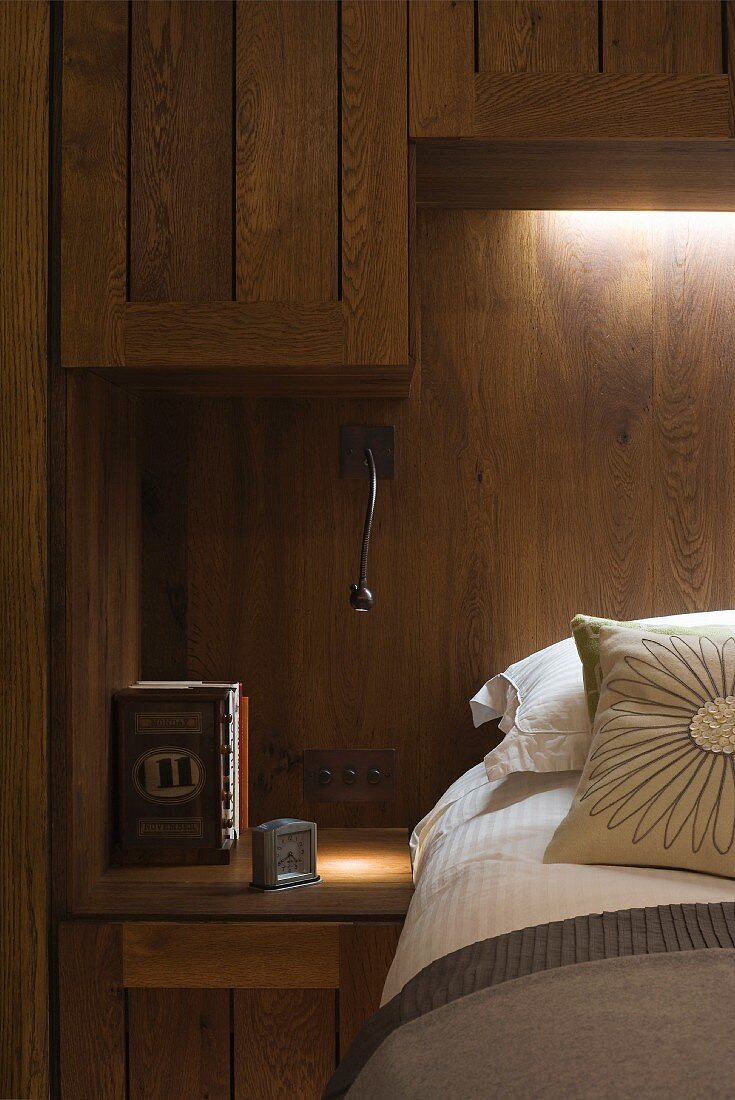 Bett vor holzverkleideter Wand mit indirekter Beleuchtung und Hängeschränkchen aus Holz