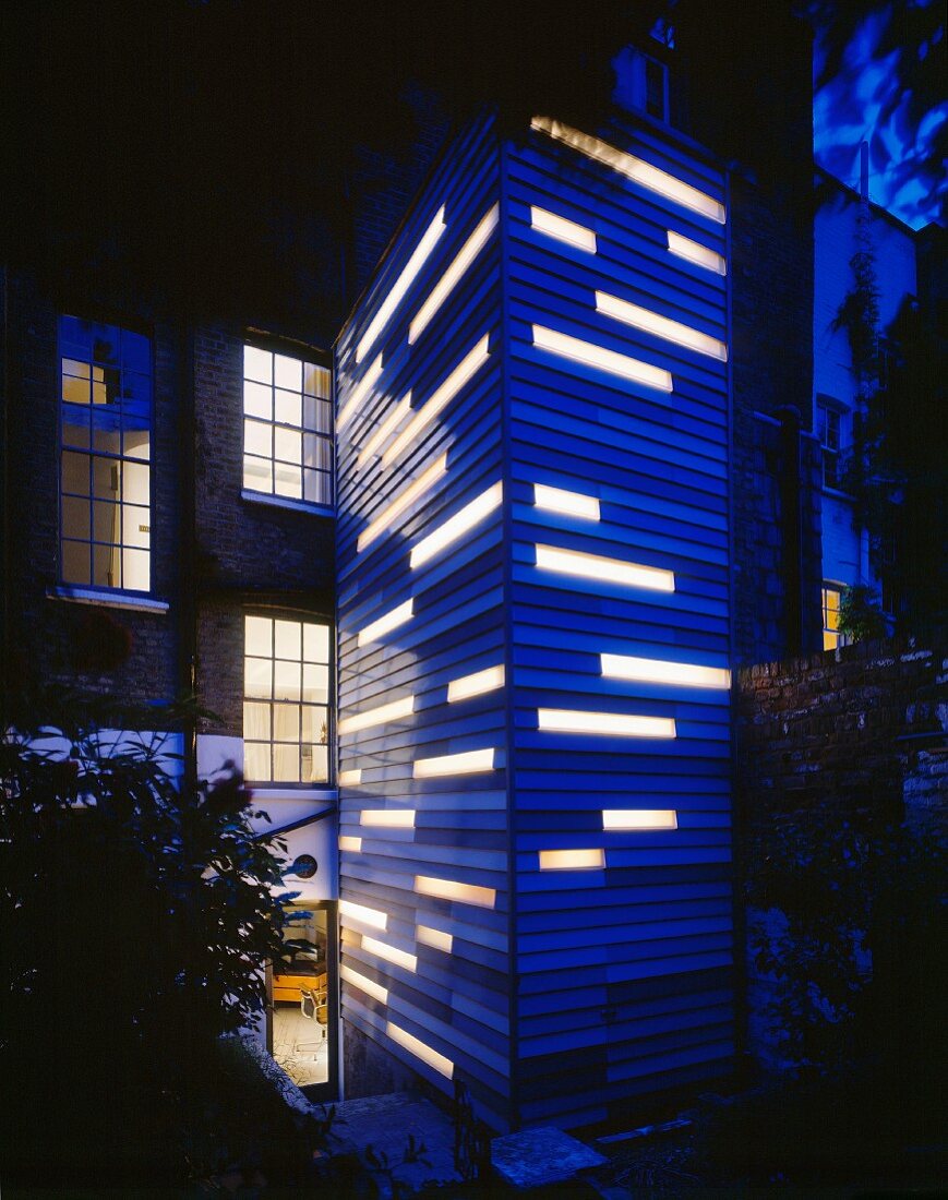 Blauer Holzverkleideter Turmanbau mit beleuchteten Fensterbändern und englisches Wohnhaus mit Ziegelfassade