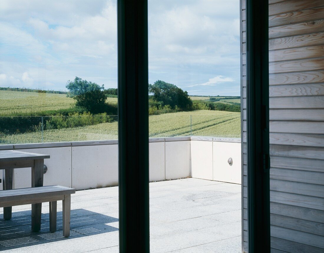 Blick durch Terrassenfenster auf Tisch und Bankgarnitur auf Terrasse vor englischer Landschaft