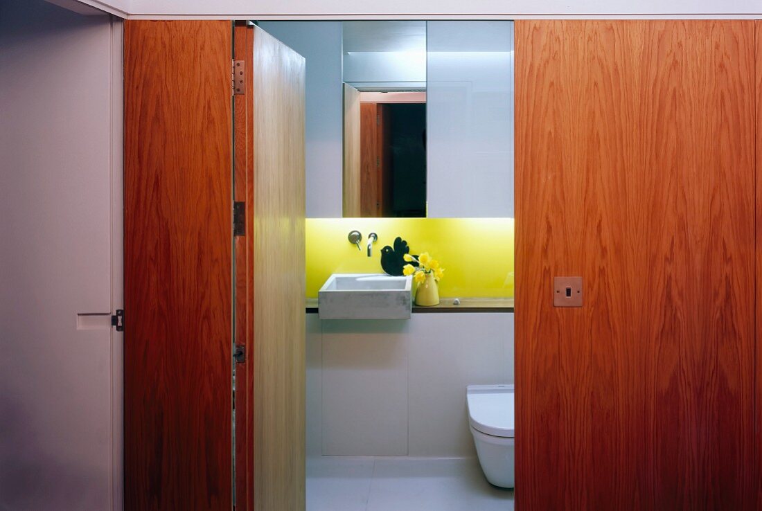 Blick durch offene Tür aus Holz in kleines designtes Bad mit Waschbecken und gelb lackierter Rückwand