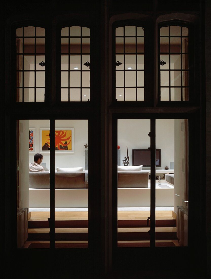 View through illuminated terrace doors with transom windows to man seated in living room