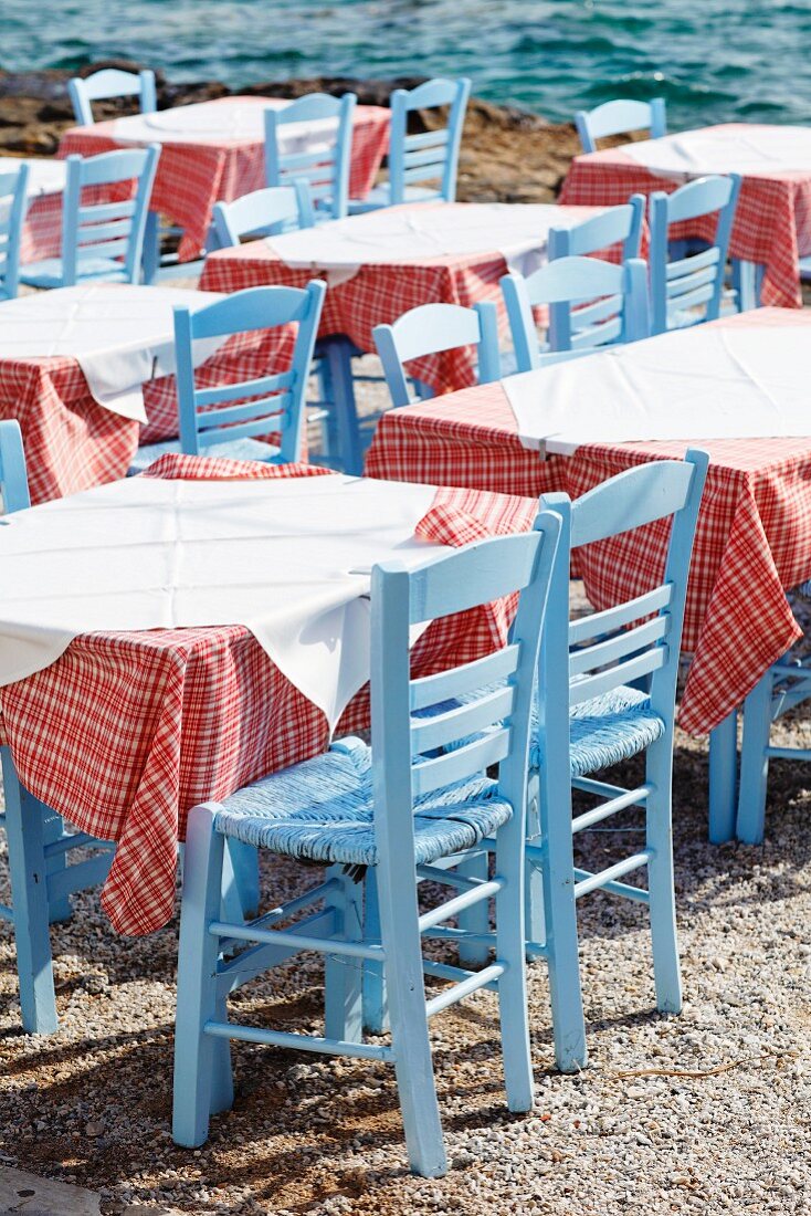 Tische mit rot-weiss karierten Tischdecken und pastellblauen Binsenstühlen in einer griechischen Taverne am Meer