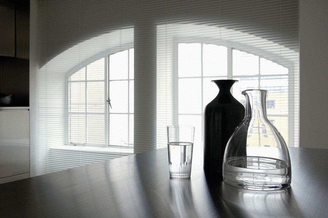 Open venetian blind in front of large split window with arched lintel, carafes and water glass in foreground