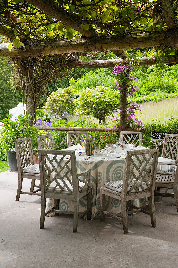 Runder Tisch mit gemusterter Tischdecke unter bewachsener Pergola im Garten