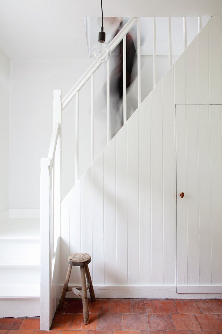 Storage space in fitted cupboard below simple, white country-house staircase; person walking down stairs and stool on terracotta floor