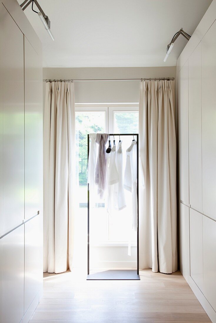 Dressing room with white fitted wardrobes and clothing on coat hangers on metal frame in front of French windows