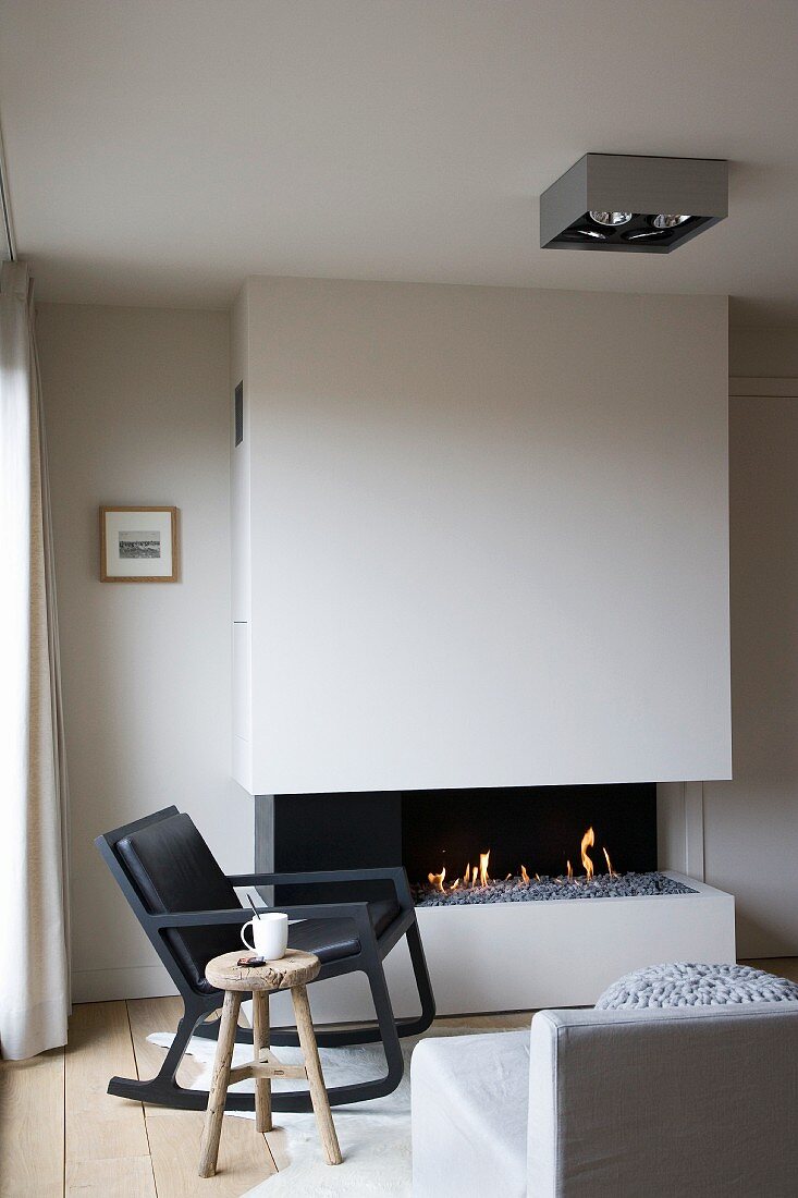 Kaminfeuerromantik im modernen Schwarzweiss-Wohnraum mit Stilmelange aus puristischem Schaukelsessel und rustikalem Holzschemel