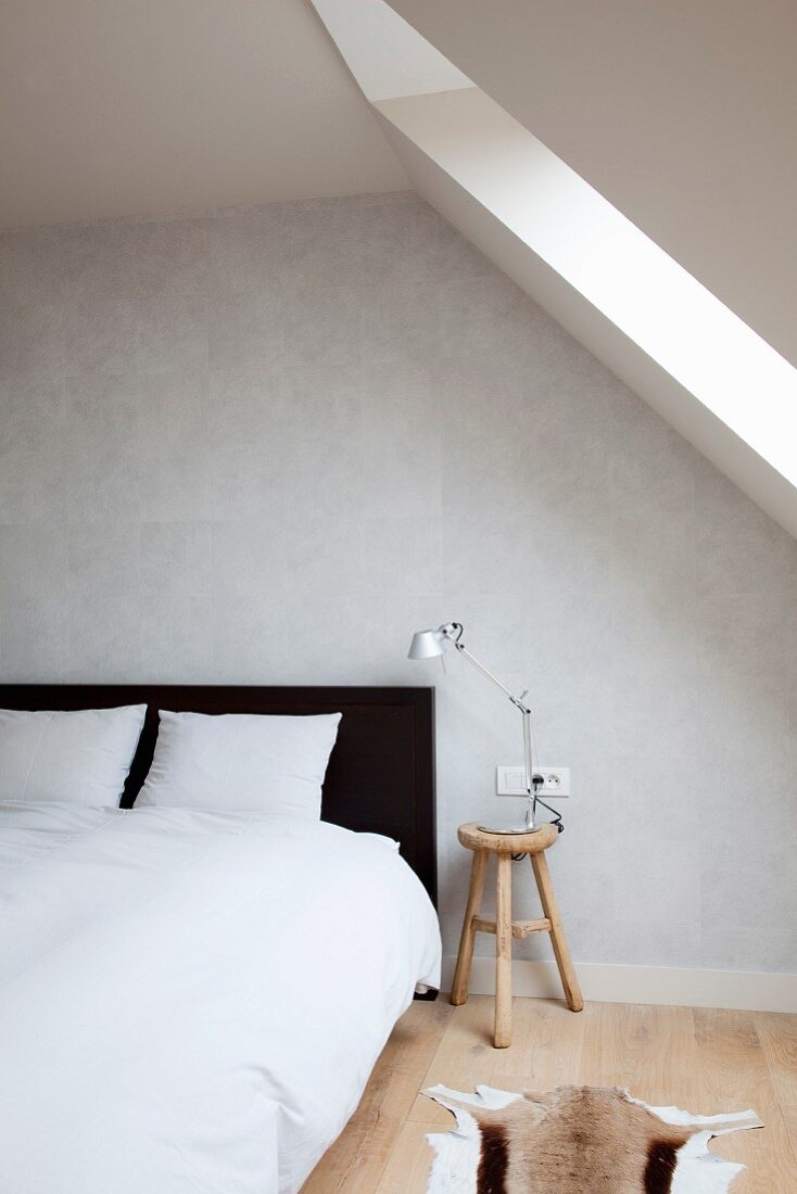 Black and white attic bedroom with double bed and grey-painted wall contrasting with warm wood of bedside stool and board floor