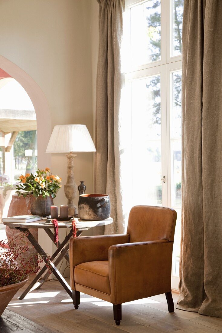 Comfortable seating area with leather armchair and side table next to French windows with floor-length curtains