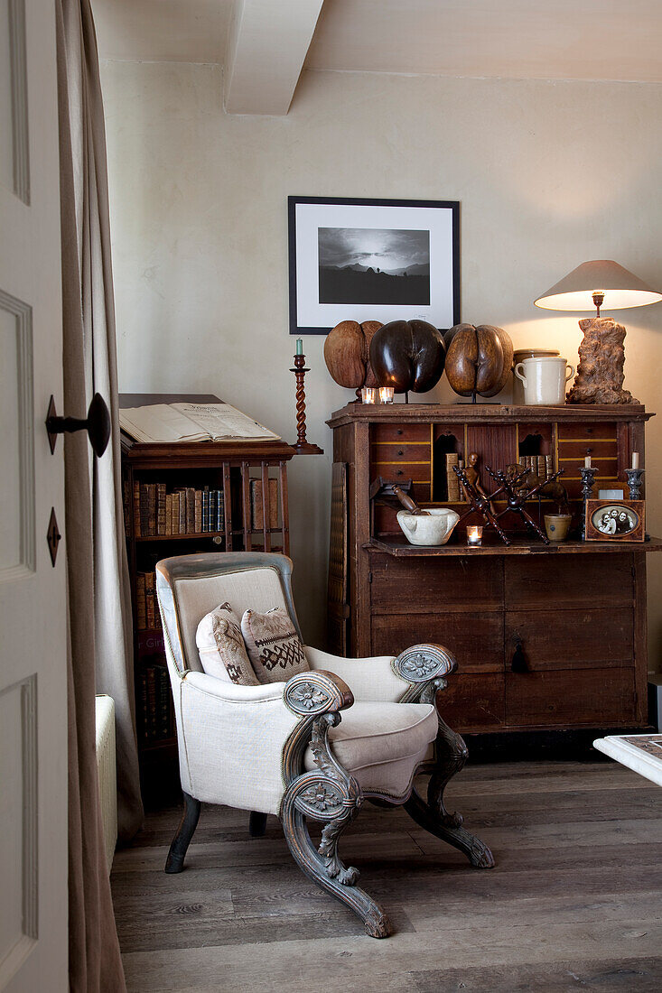 View through open door of antique armchair with white cover and carved armrests next to bureau