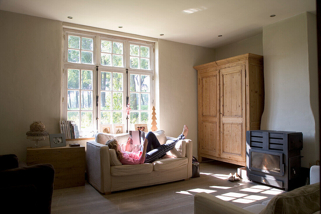 Woman reading on couch in living room with fireplace and rustic wooden cabinet