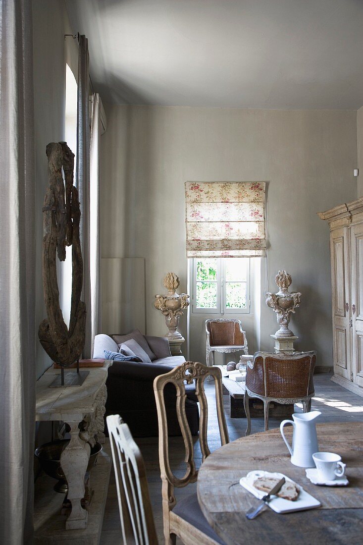 Stühle mit geschwungener Lehne an rustikalem Esstisch mit Vintage Frühstücksgeschirr in offenem Wohnzimmer mit Salonecke und Rokokostühlchen