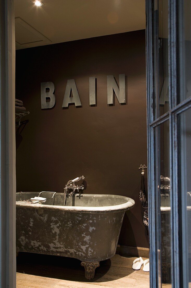 View of vintage bathtub with feet below lettering on wall painted dark brown through open glass door with metal frame