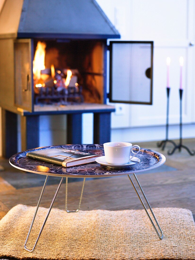 Gemütliche Winterstimmung mit brennendem Feuer im Kaminofen; davor ein runder Tabletttisch mit einer Tasse Kaffee und einem Buch