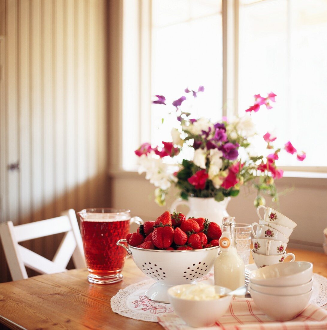 Sommerlicher Tisch mit buntem Blumenstrauss, Erdbeeren, Sahne und Fruchtsaft in einer Glaskaraffe
