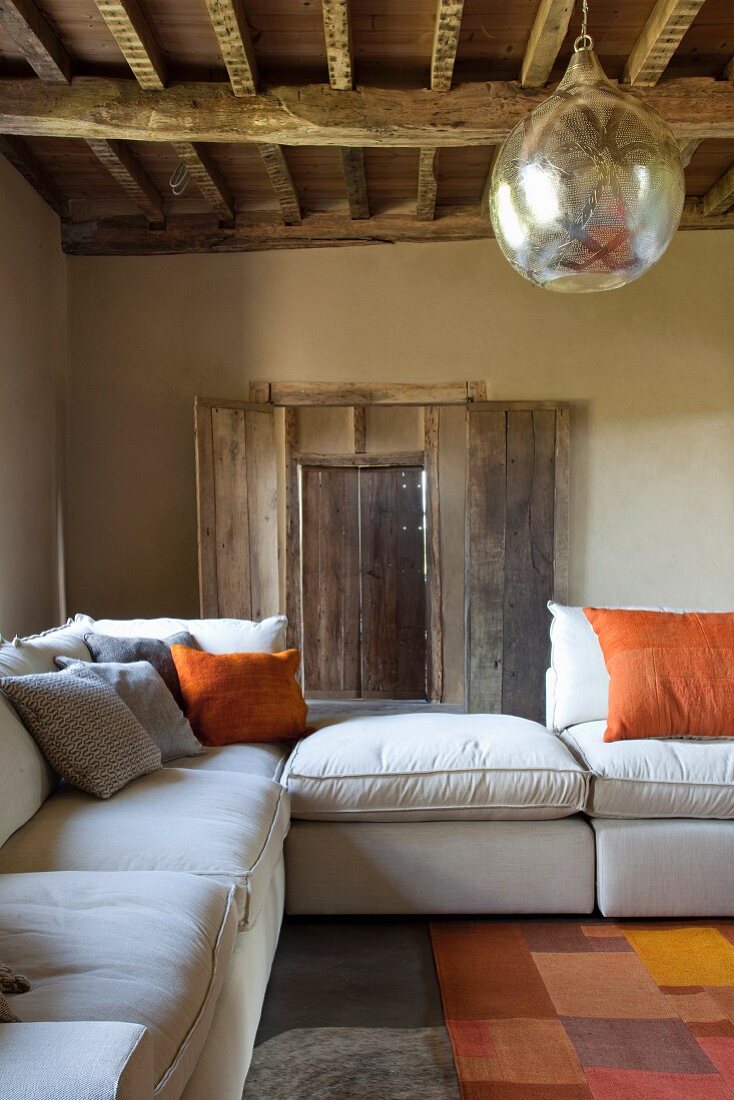 Pale sofa set and closed rough window shutters in rustic country house with spherical silver lamp hanging from wood-beamed ceiling