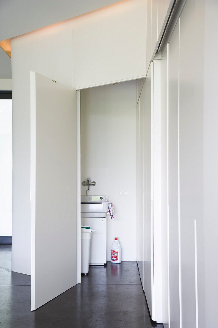 Smooth, white doors of fitted cupboards and dark floor with view of kitchen sink through open door