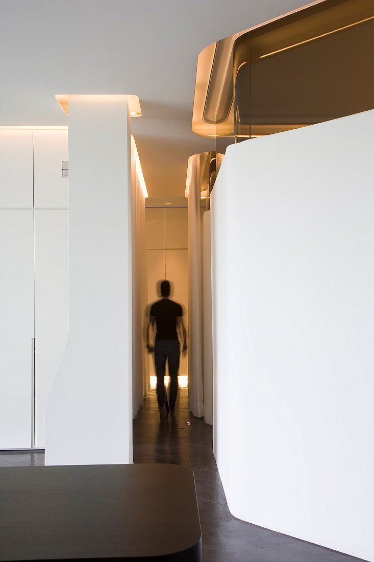 View along narrow corridor with silhouette of walking person, indirect lighting through ceiling slots running along walls