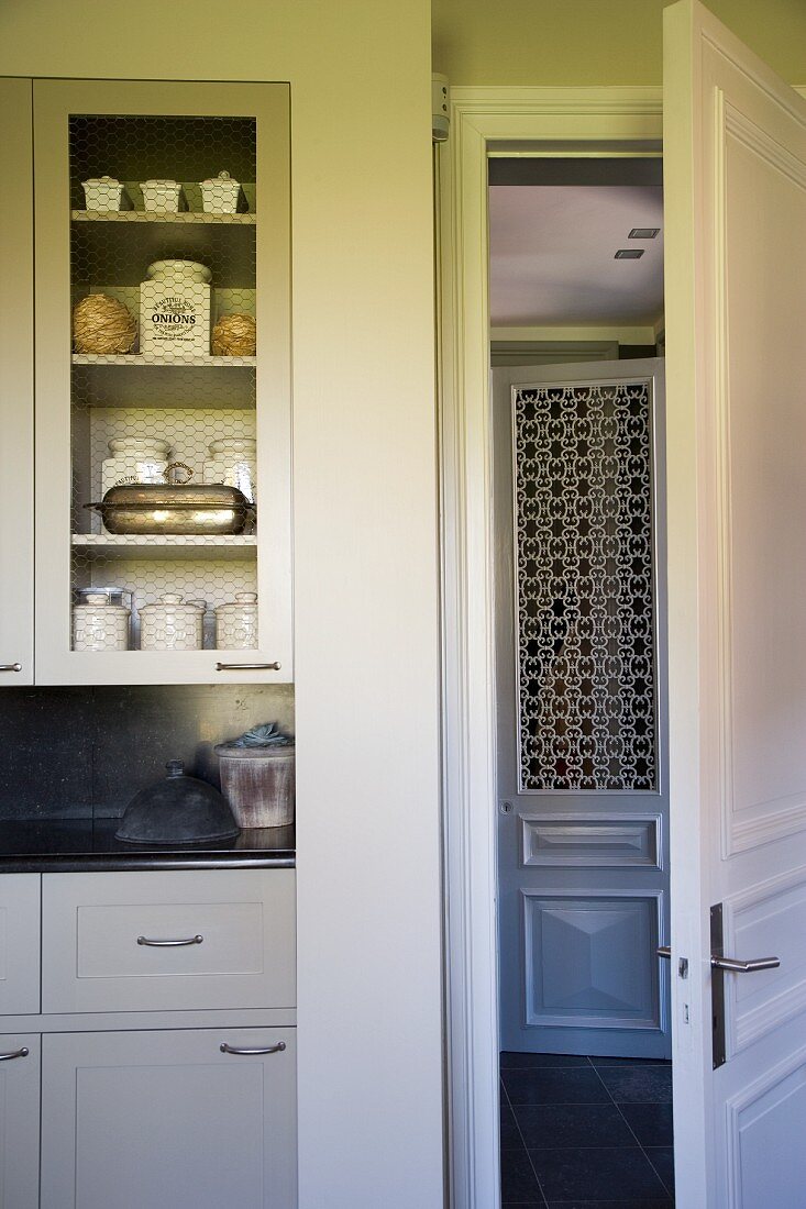Moderne, weiße Landhausküche mit Porzellangefässen im Retrostil, sichtbar hinter Gittertür und Blick auf den gemusterten Glaseinsatz einer Zimmertür