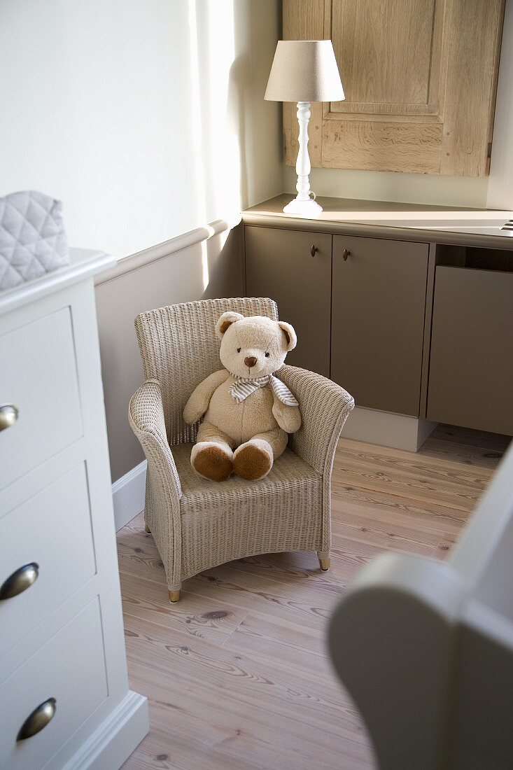 Teddybär auf Kinder-Korbstuhl zwischen gedrechseltem Lampenfuss und weisser Landhaus-Kommode