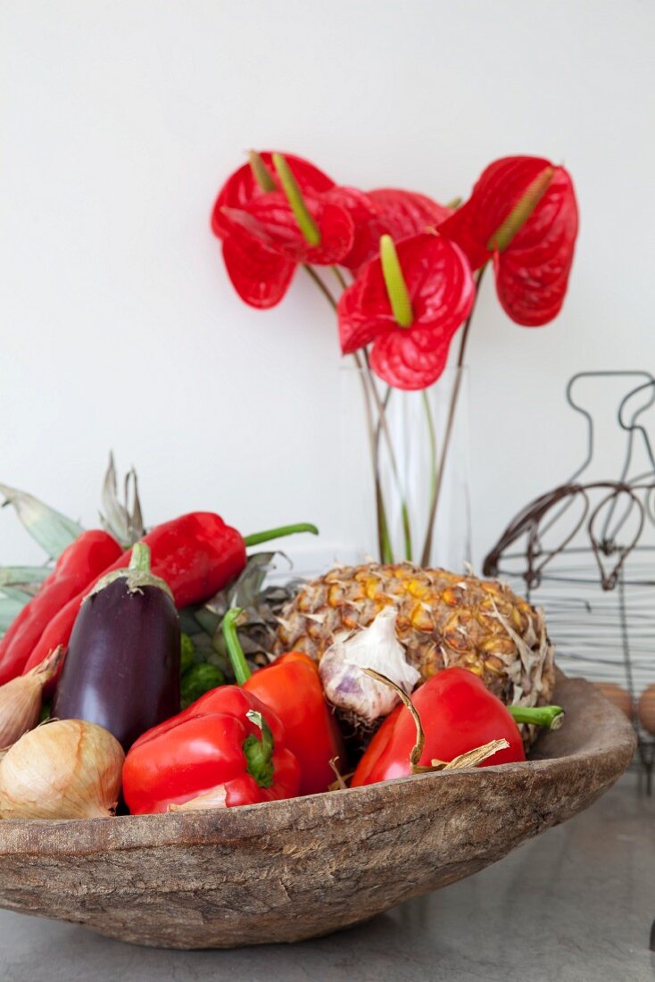 Fruit and vegetables in rustic bowl in front of glass vase of tropical flowers