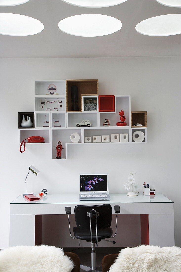Cubic desk, black swivel chair and combination of wall-mounted shelving units below circular light recesses in ceiling