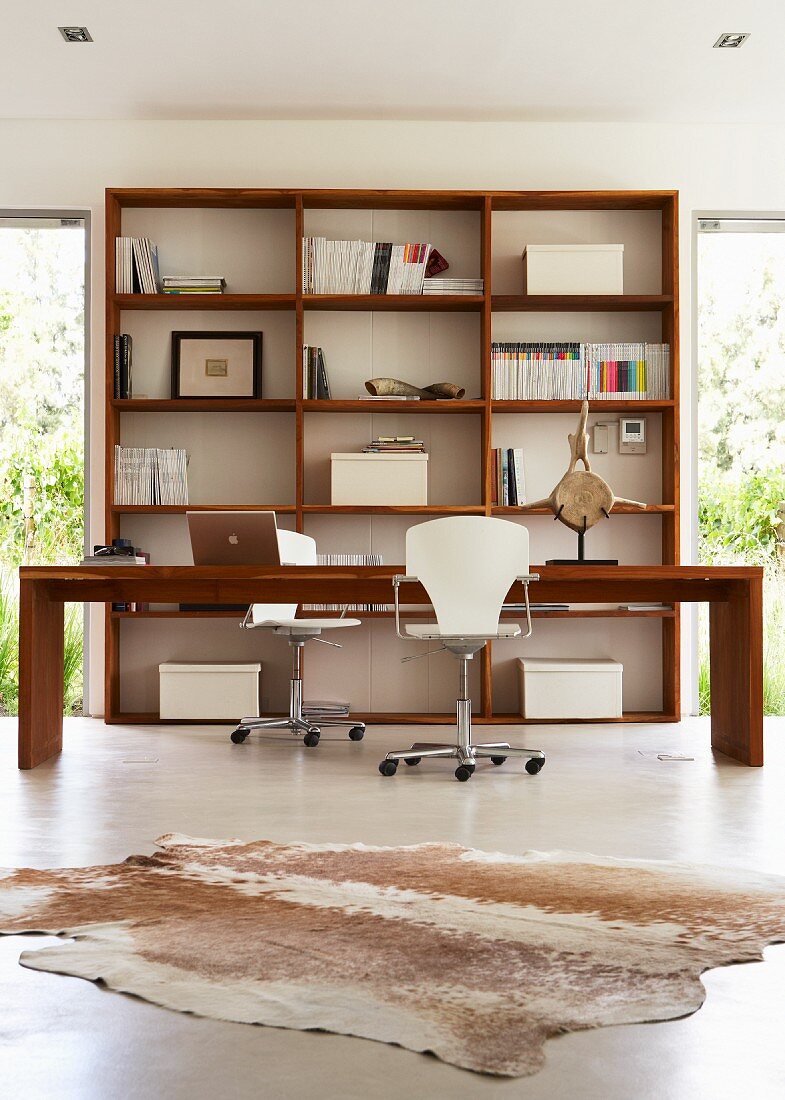 Office area flooded with light with long desk and two swivel chairs in front of open wooden shelving