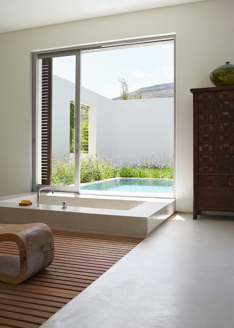 Modernes, exotisches Luxusbad - in den Boden eingelassene Wanne vor einem offenen Schiebefenster mit Blick auf einen Innenhof mit Pool