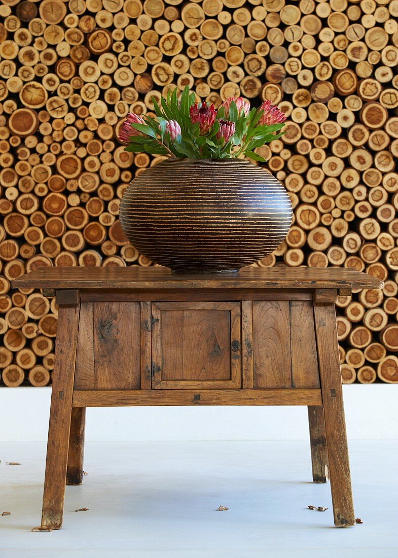 Bauchige, braungestreifte Vase mit exotischen Protea-Blüten auf altem Holzkommodentisch und sauber gestapeltes Brennholz im Hintergrund