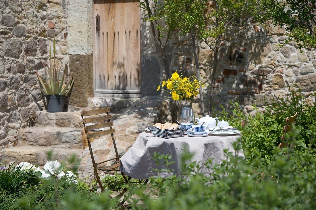 Gedeckter Frühstückstisch im Garten mit gelbem Blumenstrauss vor mediterranem Natursteinhaus