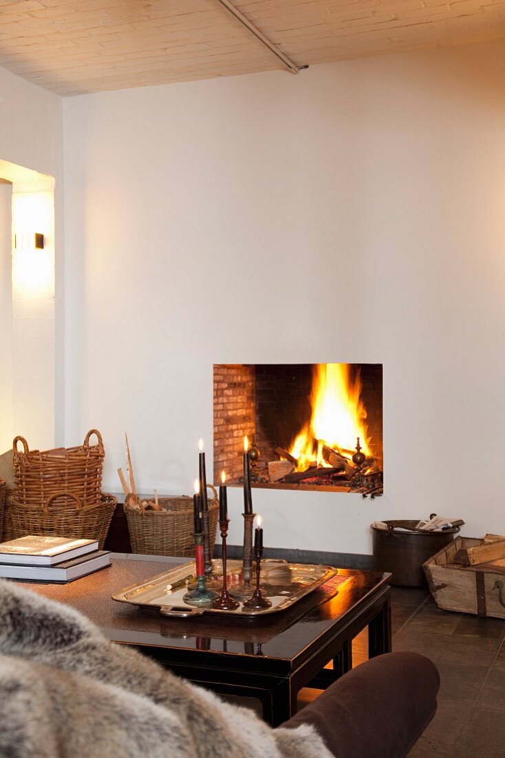 Sofa and candles on table in front of roaring fire in simple fireplace opening in smooth white wall