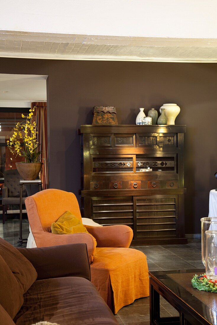 Dunkles Wohnzimmer mit antikem Holzschrank vor brauner Wand und orangefarbenem Hussensessel als Farbkontrast