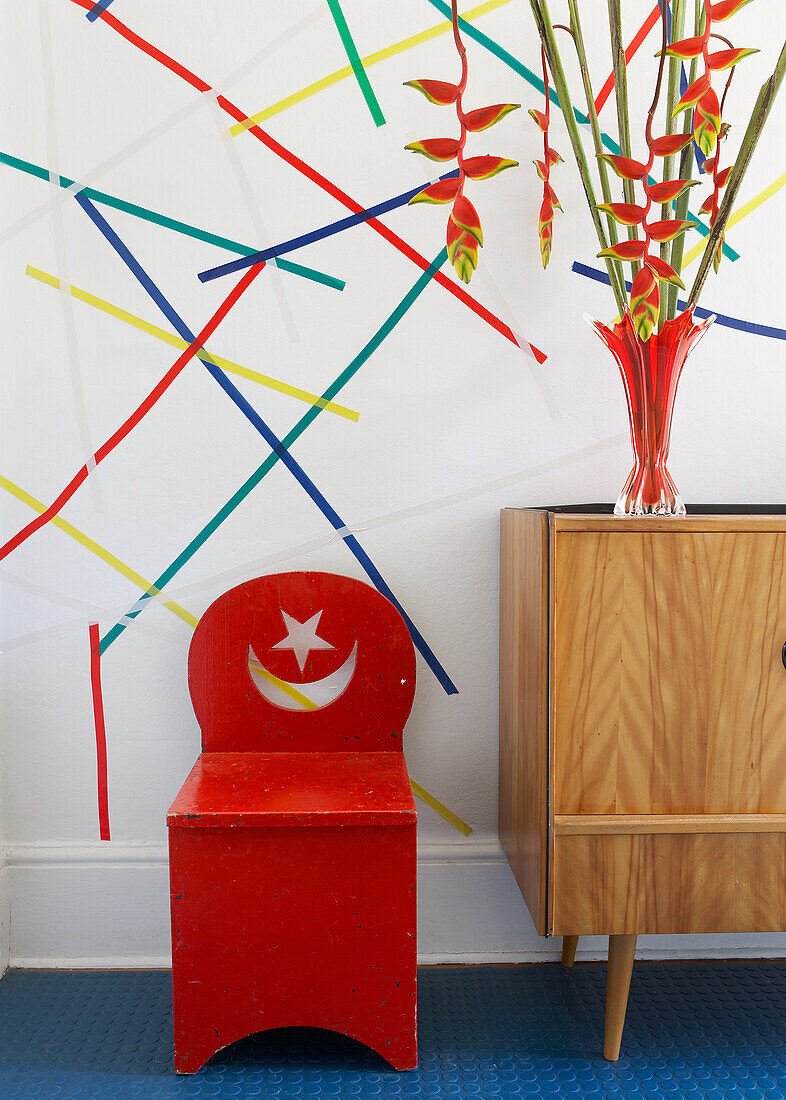 Red chair and chest of drawers in front of wall with colourful tape art pattern