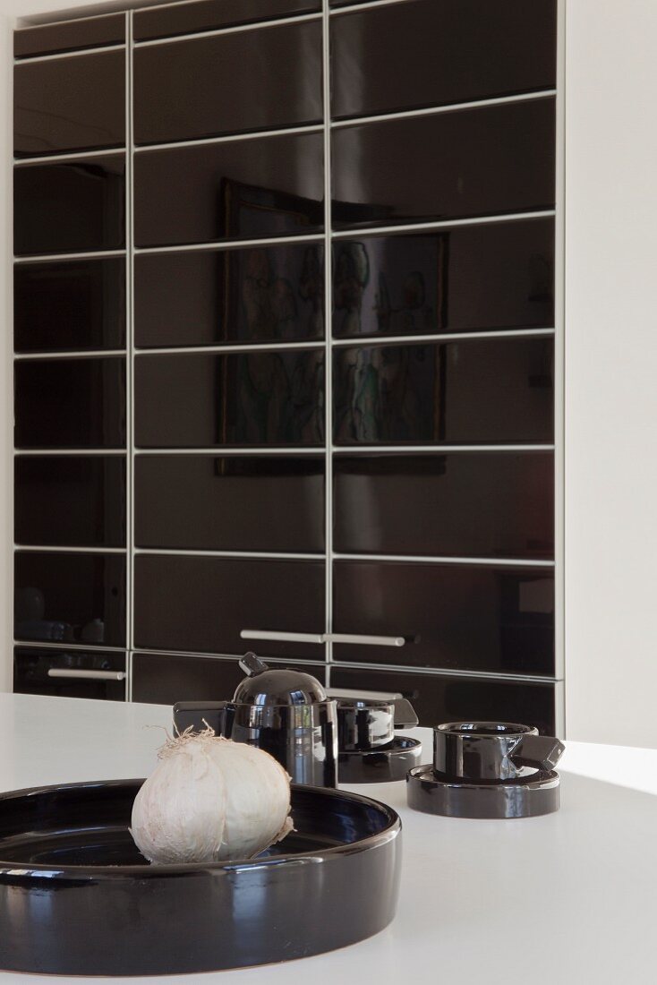 Schwarze Küchenutensilien mit weisser Knoblauchknolle vor einem Wandschrank mit fliesenartigen Türfronten