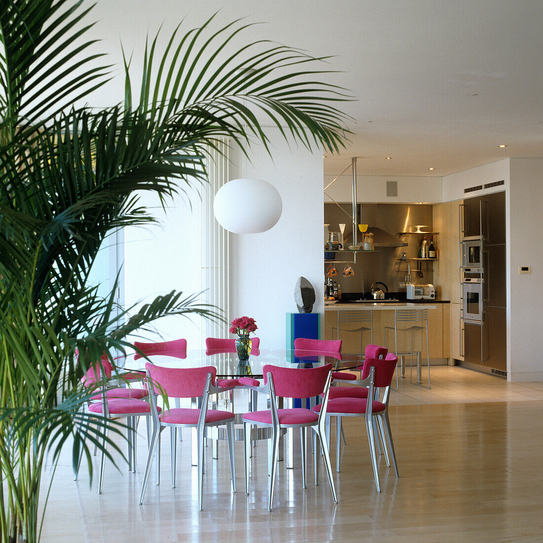 Dining area with pink chairs and palm tree, open kitchen in the background