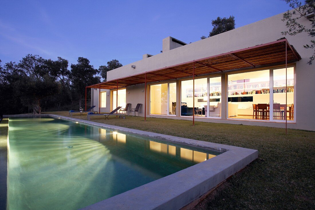 Hell erleuchtete Innenräume eines modernen Landhauses mit großem Swimmingpool und sich spiegelndem Licht